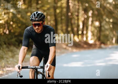 Bärtiger Mann mit sportlichem Körper trägt Sportkleidung, Schutzhelm und Sonnenbrille Reiten schwarzen Fahrrad auf gepflasterter Straße mit verschwommenem Hintergrund. Konzept Stockfoto