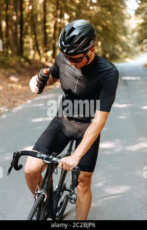 Radprofi in Schutzhelm und Brille hielt im grünen Wald, um frisches Wasser zu trinken. Reifer Mann in activewear Einnahme Ruhe während RID Stockfoto