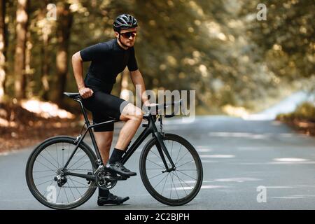 Sportlicher bärtiger Mann in Radbekleidung, Schutzhelm und Spiegelbrille posiert auf Kamera mit schwarzem Fahrrad im Freien. Konzept von Sport und aktiv l Stockfoto