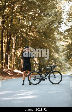 Selbstbewusster bärtiger Radfahrer im besonderen Sport-Outfit, der neben seinem schwarzen Fahrrad steht und die Kamera anschaut. Reifer Mann in Schutzhelm und verspiegelte gla Stockfoto