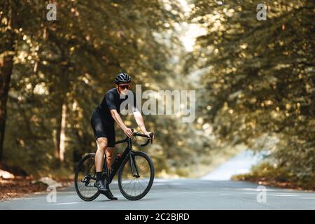 Reife Sportler mit sportlicher Körperform in Radbekleidung, Helm und Brille stehend mit Fahrrad im Sommerwald gekleidet. Bärtiger Mann genießt fre Stockfoto