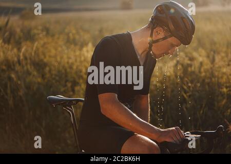 Seitenansicht eines reifen Radfahrers mit müdem nassen Gesicht, das schwarzen Helm trägt und Sportkleidung, die auf dem Fahrrad im Freien sitzt. Bärtiger Mann, der sich ausruhte Stockfoto