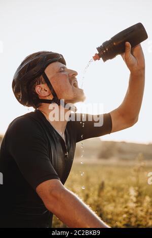 Seitenansicht des erschöpften bärtigen Sportlers in schwarzem Helm, der Wasser aus der Sportflasche auf sein Gesicht gießt. Reifer Mann erfrischend nach langen Strecke Reiten Stockfoto