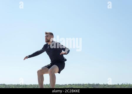 Porträt eines jungen bärtigen Athleten von 25 Jahren, der gegen einen blauen Himmel Athleten springt, der entlang der Küste läuft, Draufsicht. Aktiver Lebensstil. Junger Mann Stockfoto