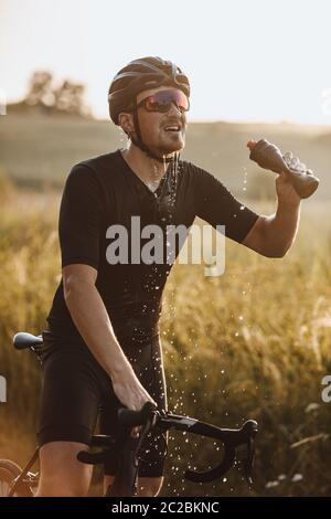 Starker bärtiger Mann in aktiver Kleidung, Schutzhelm und Sportbrille spritzt frisches Wasser aus der Flasche im Freien. Müde Radfahrer nach Durst Stockfoto