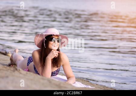 Die lächelnde Sonnenfrau genießt es im Sommer, sich auf Sand am Strand zu legen. Stockfoto