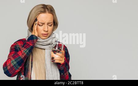 Krank junge blonde Frau in kariertem Hemd, gewickelter Schal mit Kopfschmerzen und Glas mit Wasser halten, berühren ihren Tempel und geschlossenen Augen, Nahaufnahme Stockfoto