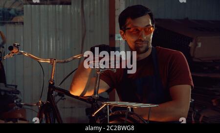 Thema kleine Unternehmen Fahrrad Reparatur. Ein junger kaukasischer Brünette Mann trägt Schutzbrille, Handschuhe und eine Schürze verwendet ein Handwerkzeug Stockfoto