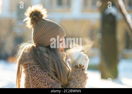 Nette junge kaukasische Frau in beigem Pelzmantel, Hut mit Pompon, Schal und weißen Fäustlingen halten dampfende weiße Tasse heißen Tee oder Kaffee, im Freien in der Sonne Stockfoto