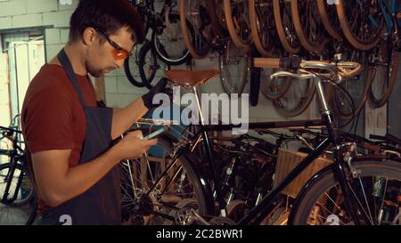 Thema kleine Unternehmen Fahrrad Reparatur. Junge kaukasische Brünette Mann trägt Schutzbrille, Handschuhe und fartukhe nutzt Handy tec Stockfoto