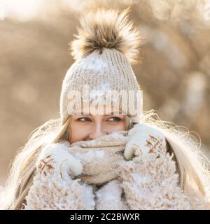 Portrait einer hübschen jungen Frau mit langen Haaren in beigem Hut mit Pelzpompon, warmem Schal, Mantel, weißen Handschuhen mit Schnee bedeckt an sonnigen Wintertag. Fema Stockfoto
