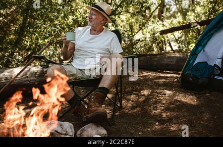Rentner, der am Lagerfeuer saß und Kaffee getrunken hat. Ältere männliche Camping im Wald mit einem erfrischenden Kaffee am Morgen. Stockfoto
