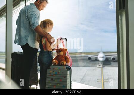 Vater und Tochter schauten Flugzeuge an, während sie auf dem Flughafen warteten. Mann mit seiner Tochter, die an einem großen Fenster am Flughafenterminal steht. Stockfoto