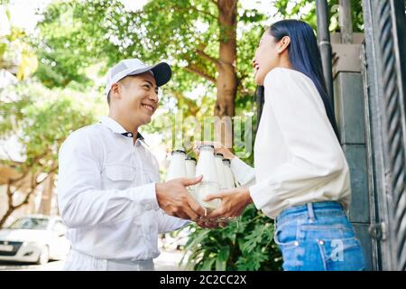 Horizontale Low-Winkel-Aufnahme von fröhlichen Mann trägt weiße Uniform Lieferung Milchflaschen an glückliche junge Frau Stockfoto