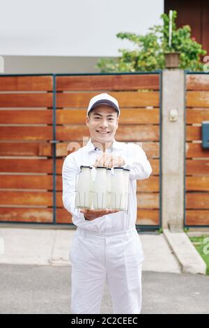 Schöner asiatischer Milchmann trägt perfekte weiße Uniform steht gegen Holzzaun hält Glasflaschen mit Milch, vertikal Medium lang Schuss, Kopie sp Stockfoto