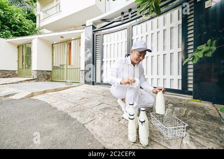 Fröhlicher asiatischer Milchmann trägt perfekte weiße Uniform, Milch abliefern und leere Flaschen am frühen Morgen zurücknehmen, Platz kopieren Stockfoto