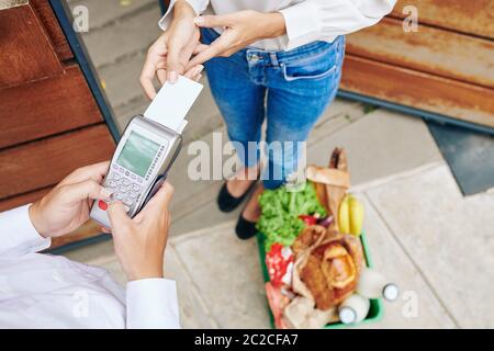 Horizontale Aufnahme von oben, in der eine unerkennbare junge Frau mit ihrer Debitkarte für den Lieferservice von frischen Lebensmitteln bezahlt Stockfoto