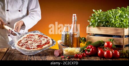 Koch, eine traditionelle italienische Pizza in der Pizzeria Holding eine Torte mit Tomate und Käse auf einem Paddel mit verschiedenen frischen Zutaten über eine Bro Stockfoto