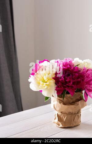 Schöner Blumenstrauß von Pfingstrosen steht auf einem weißen Tisch vor einem Hintergrund von grauen Vorhängen und einer weißen Wand Stockfoto