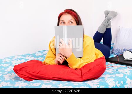 Junge schöne Mädchen auf dem Bett in einem Schlafzimmer, neben einem Laptop und Kopfhörer, die ein Buch über ihr Gesicht und versuchte, zu studieren, sondern sie ist Stockfoto