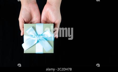 Weibliche Hände halten ein kleines Geschenk mit blauem Band auf schwarzem Hintergrund verpackt Stockfoto