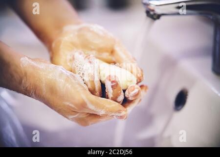 Ein Mann wäscht sich vorsichtig mit einem weißen Stück Seife über dem Waschbecken, in das ein Wasserstrom aus dem Wasserhahn fließt. Stockfoto