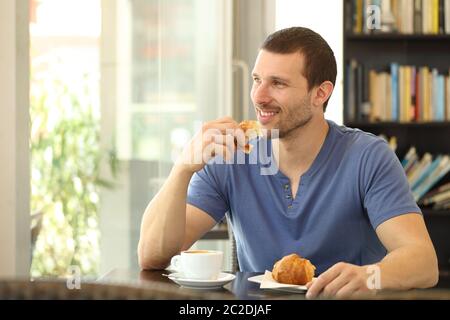 Ein glücklicher Mann, der ein Croissant zum Frühstück isst und in einem Café sitzt und durch ein Fenster schaut Stockfoto