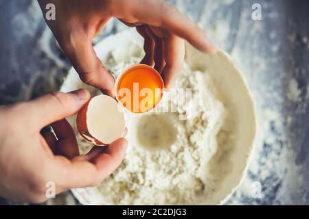 Der Koch hält ein gebrochenes Hühnereier mit dem Eigelb in der Schale und wird es mit Mehl in einer weißen Schüssel mischen. Backvorgang für Zuhause. Stockfoto