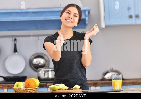 Eine junge asiatische Frau in einem schwarzen T-Shirt macht eine Frucht Frühstück und schneidet Apple auf einem Holzbrett in einer Küche. Orangensaft und Obst in der Schüssel ist Stockfoto