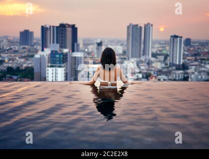 Junge Frau entspannen Sie sich im Swimmingpool auf dem Dach während des atemberaubenden Sonnenuntergangs und genießen Sie den Blick auf die Stadt Stockfoto
