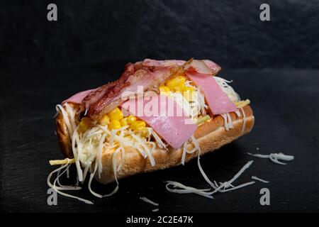 Venezolanischer Hot Dog mit geriebenem Käse, Mais, Speck und süßem Schinken auf schwarzem Hintergrund. Stockfoto