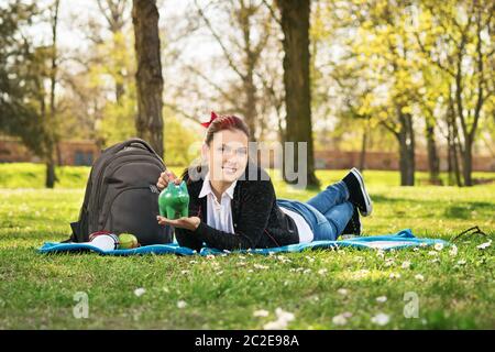 An meine Zukunft und Speichern mit Leichtigkeit. Schöne junge Studentin liegen auf einer Wiese im Park, ein Sparschwein. Stockfoto