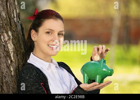 Speichern während der sonnigen Tage so, wenn die Regenzeit kommt. Schöne junge Schülerin lehnte sich an einen Baum im Park, eine Münze in ein Sparschwein. Stockfoto