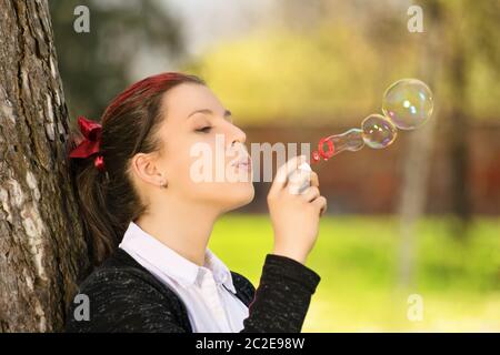 Eine Nahaufnahme portrait Schuß eines schönen jungen Schüler Mädchen in einem Park, lehnte sich an einen Baum, bläst Seifenblasen. Stockfoto