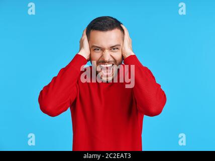 Gestresster emotionaler junger Mann schreit und schließt Ohren durch seine Hände isoliert auf blauem Hintergrund Stockfoto