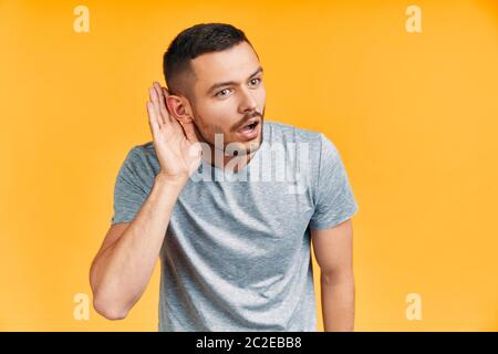 Der junge überraschte Mann hört etwas vorsichtig zu und hält seine Hand nahe am Ohr über gelben Hintergrund Stockfoto