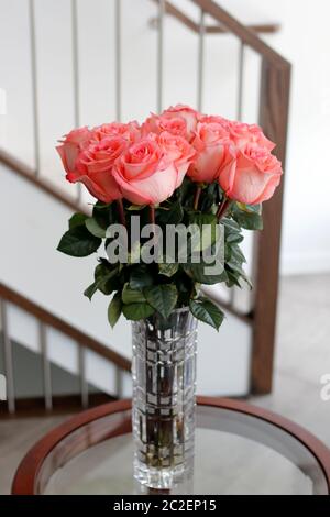 Schöne dunklere rosa Rosenblüten mit dunkelgrünen Blättern in einer Glasvase auf einem Wohnzimmertisch. Rosa Rosen Blumenstrauß in einem klaren ta Stockfoto