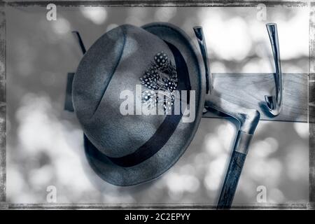 Arty, monochrome Nahaufnahme der 40er Jahre, Vintage Trilby Hut der 1950er Jahre, Fedora Hut und altmodischer Gehstock, der in gedappltem Sonnenlicht auf der Garderobe aufgehängt wird. Stockfoto