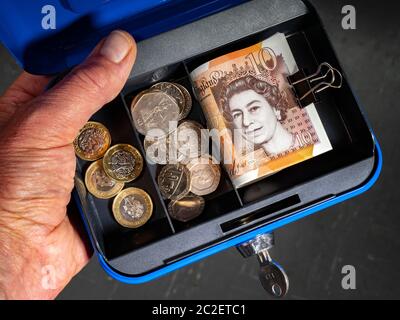 Hand des Mannes auf einem offenen kleinen, blauen, persönlichen Sicherheitskasten, mit Schlüssel im Schloss, mit einer Reihe von Sterling Pfund Bargeld, Banknoten und Münzen. Stockfoto