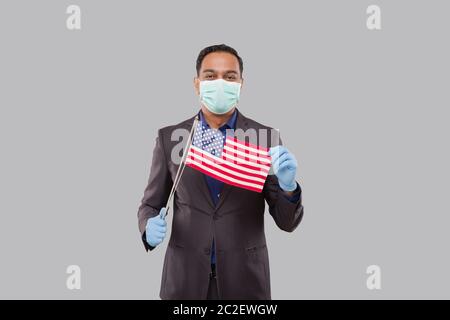 Geschäftsmann mit amerikanischer Flagge trägt medizinische Maske und Handschuhe. Indischer Geschäftsmann steht in voller Länge mit USA Flagge in Händen Stockfoto