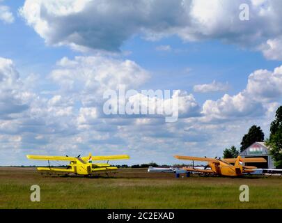 Doppelflügelige gelbe Doppeldecker. Ländlicher grasbewachsener Flugplatz. Startbereit. Segelflugplatz. Abschleppflugzeuge. Hellblauer Himmel mit dynamischen weißen Wolken Stockfoto