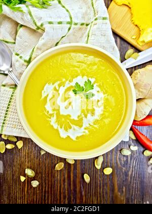 Suppe - Kürbis pürieren, mit Sahne und Petersilie in einer Schüssel auf dem Handtuch mit einem Löffel, Hülsen der heißen roten Pfeffer, Samen und Kürbis auf Holzbrett Hintergrund von einem Stockfoto
