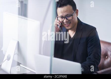 Junger asiatischer Geschäftsmann, der Handtelefon und Computer verwendet, arbeitet Stockfoto