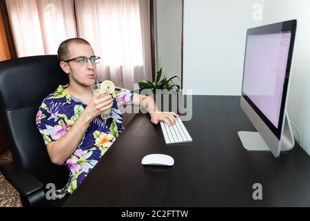 Mann, der am Computer arbeitet, während er einen erfrischenden Sommerdrink genießt und ein hawaiihemd trägt. Erfolgreicher junger Mann, Influencer arbeitet aus Stockfoto