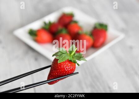 Essstäbchen halten eine frische rote Erdbeere vor einem verschwommenen Hintergrund von einem Teller Erdbeeren. Stockfoto