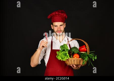 Der Koch in burgunderroter Uniform hält rote Chilischoten in der Hand. Natürliches und würziges Menü-Konzept. Mann mit Bart und Gemüse auf schwarzem Hintergrund. Kochen Sie mit lächelndem Gesicht, Chili und Korb mit frischem Gemüse. Stockfoto