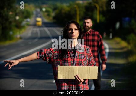 Verliebte Paare reisen durch Anhalter, kopieren Raum. Frau versuchen, Auto mit leerem Karton Zeichen und Geste zu stoppen. Reise- und Anhalter-Konzept. Paar mit nachdenklichen Gesichtern reisen mit Auto-Stopp. Stockfoto