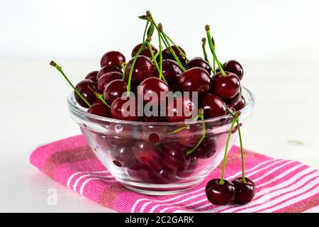 Frische Sauerkirschen in Glasschüssel, Geschirrtuch und grüne Blätter auf weißem Holztisch. Frische reife Sauerkirschen. Stockfoto