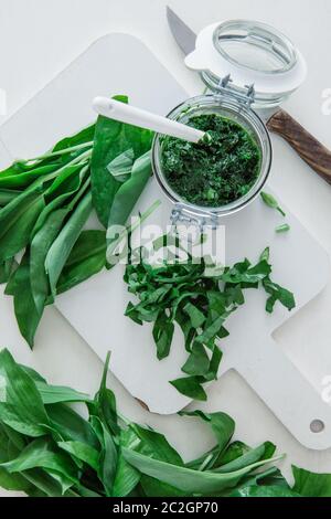 Ein Glas Pesto und grüne Blätter von Bärlauch mit Küchenutensilien auf einem weißen Tisch, Draufsicht Stockfoto