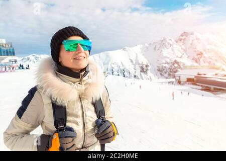 Portrait von schönen jungen Erwachsenen kaukasischen Frau in Sportanzug, Hut, Sonnenbrille und Rucksack suchen und lächeln auf Berggipfel mit Schnee bedeckt Stockfoto